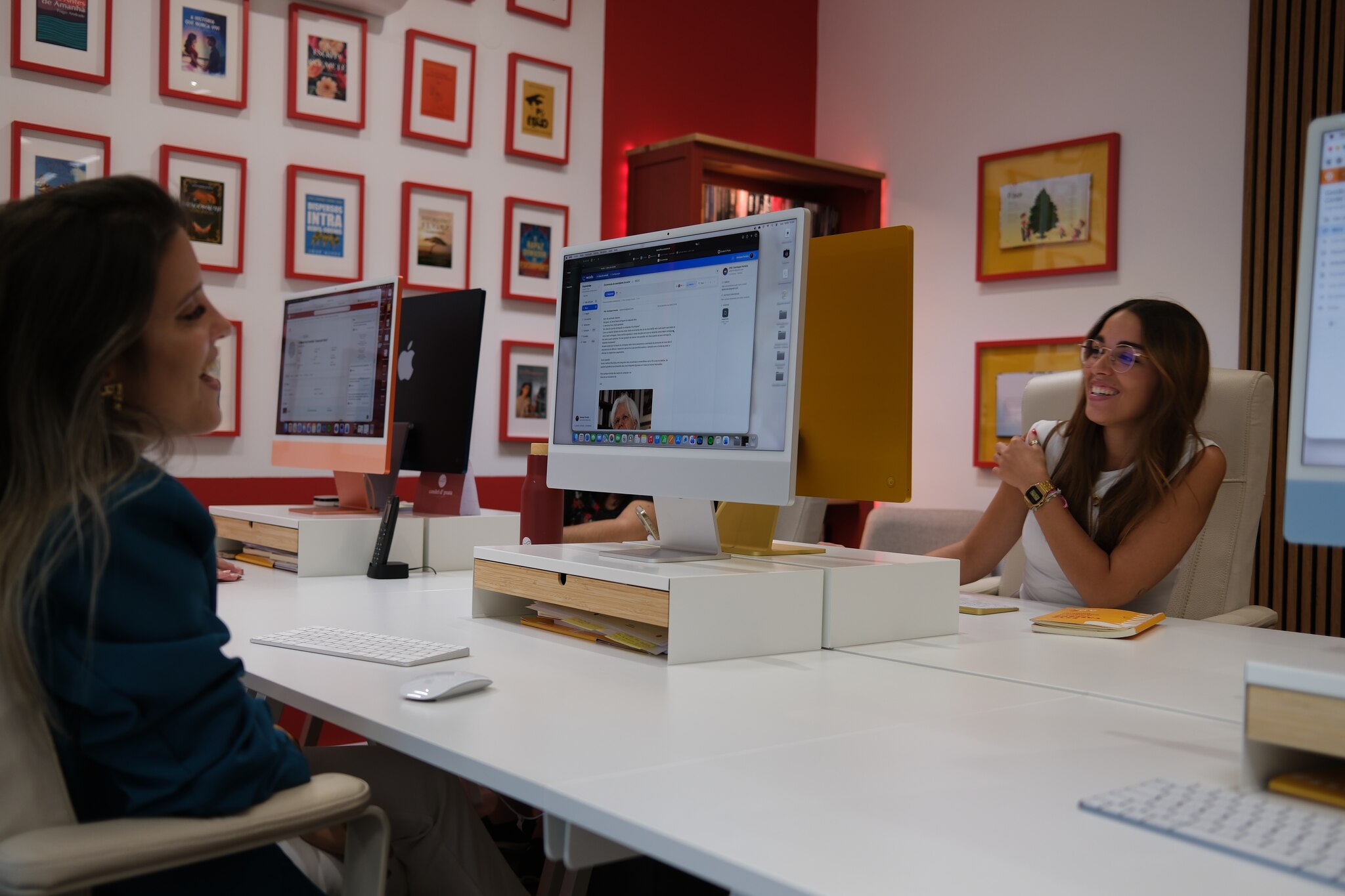 Duas pessoas da equipa aos computadores, com a parede de capas publicadas ao fundo