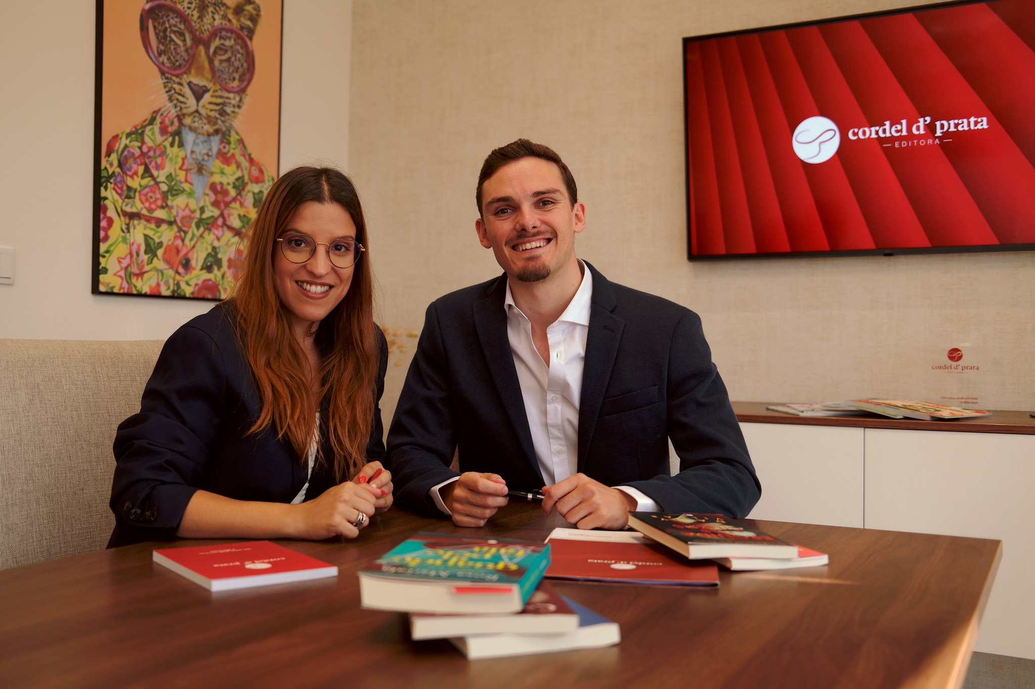Dois elementos da equipa à mesa de reunião, com livros à frente e o logótipo da editora na televisão ao fundo