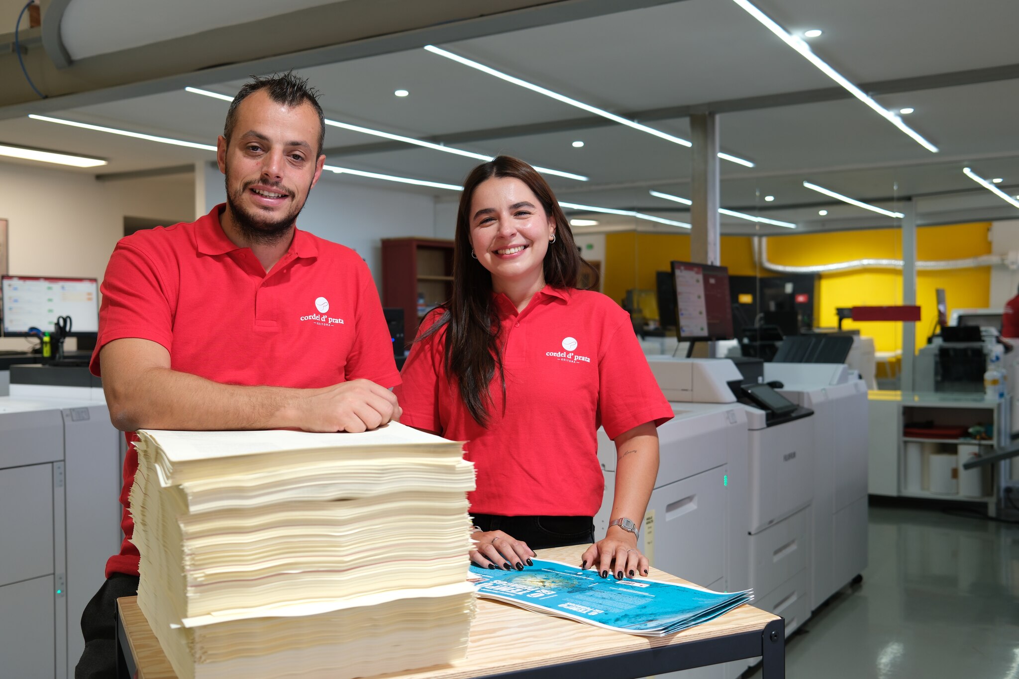Dois elementos da equipa na gráfica, com uma pilha de folhas impressas acabadas de sair da máquina