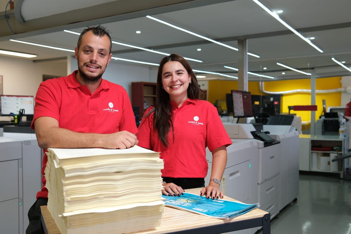 Duas pessoas da equipa, de farda vermelha, ao lado de uma pilha de folhas impressas acabadas de sair das máquinas da gráfica da Maia