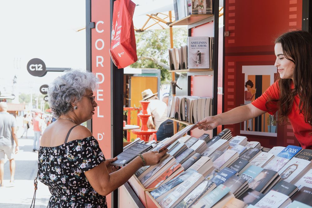 Uma visitante a folhear um livro na banca da Cordel D' Prata na Feira do Livro de Lisboa, atendida por uma colega da editora