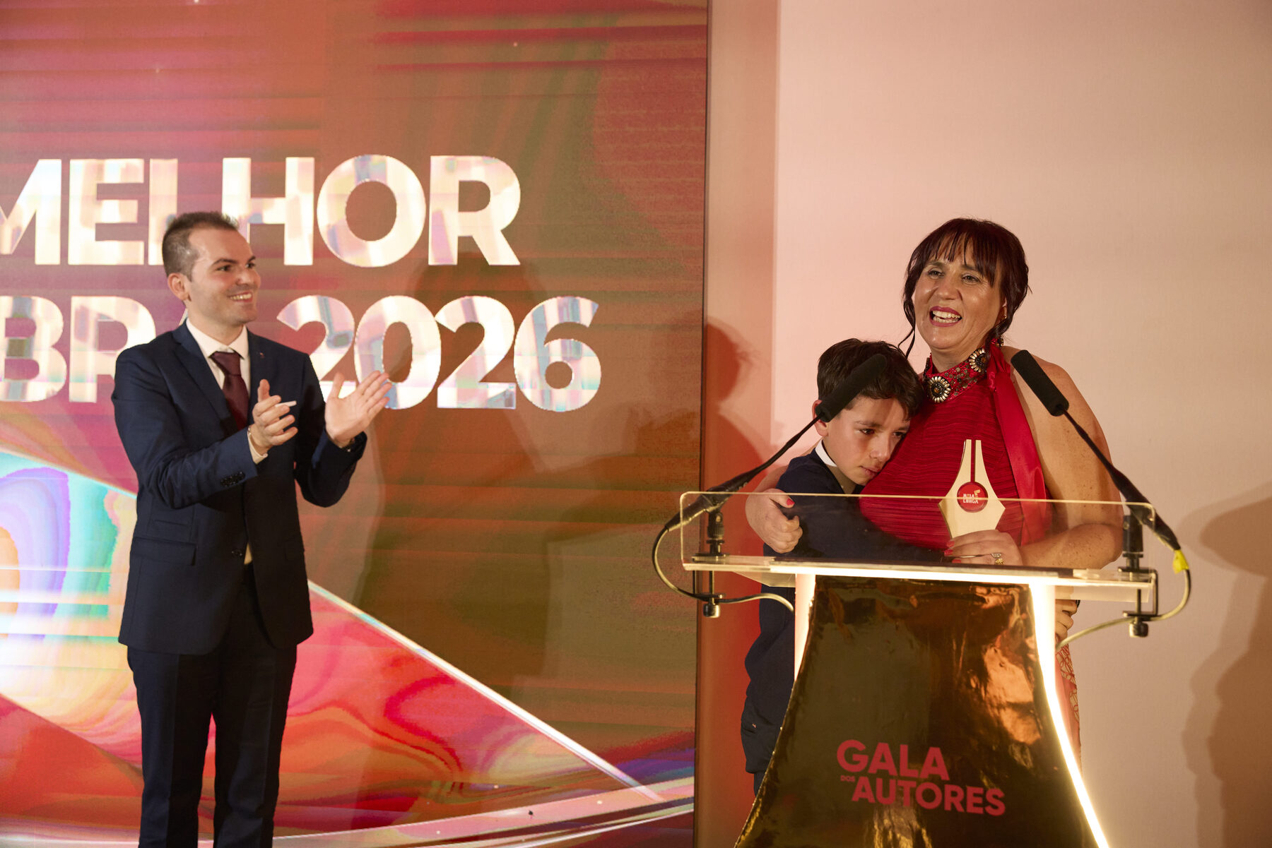 Lina Simão no palco, com o troféu e o filho abraçado a ela, diante do ecrã da Melhor Obra 2026